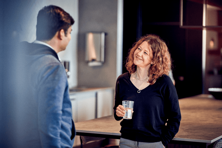 The image shows two colleagues having a friendly conversation in a casual office setting. One holds a glass of water, and both appear engaged, suggesting a relaxed and open workplace environment.