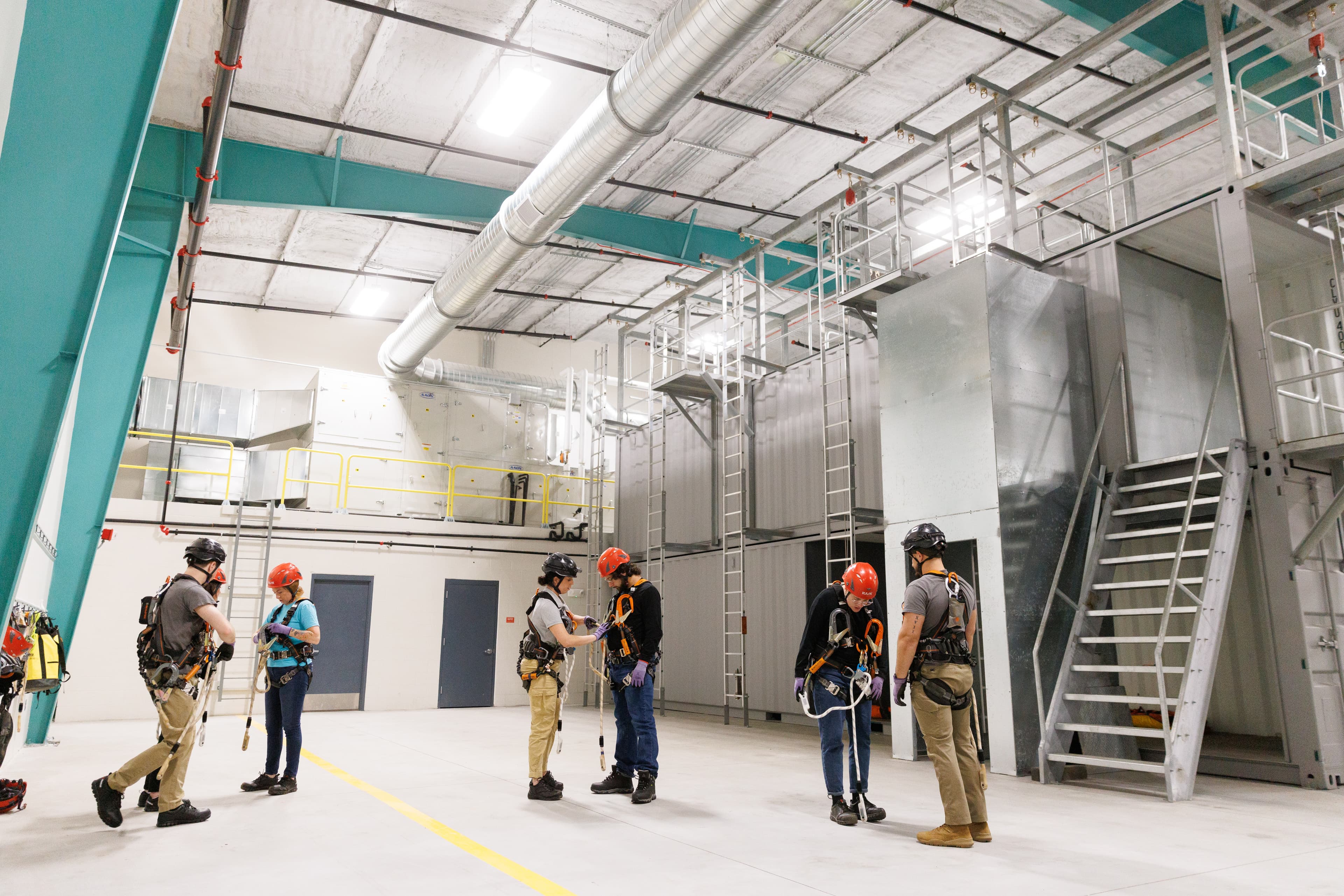 a group of individuals in safety harnesses and helmets participating in a training session within an industrial facility.